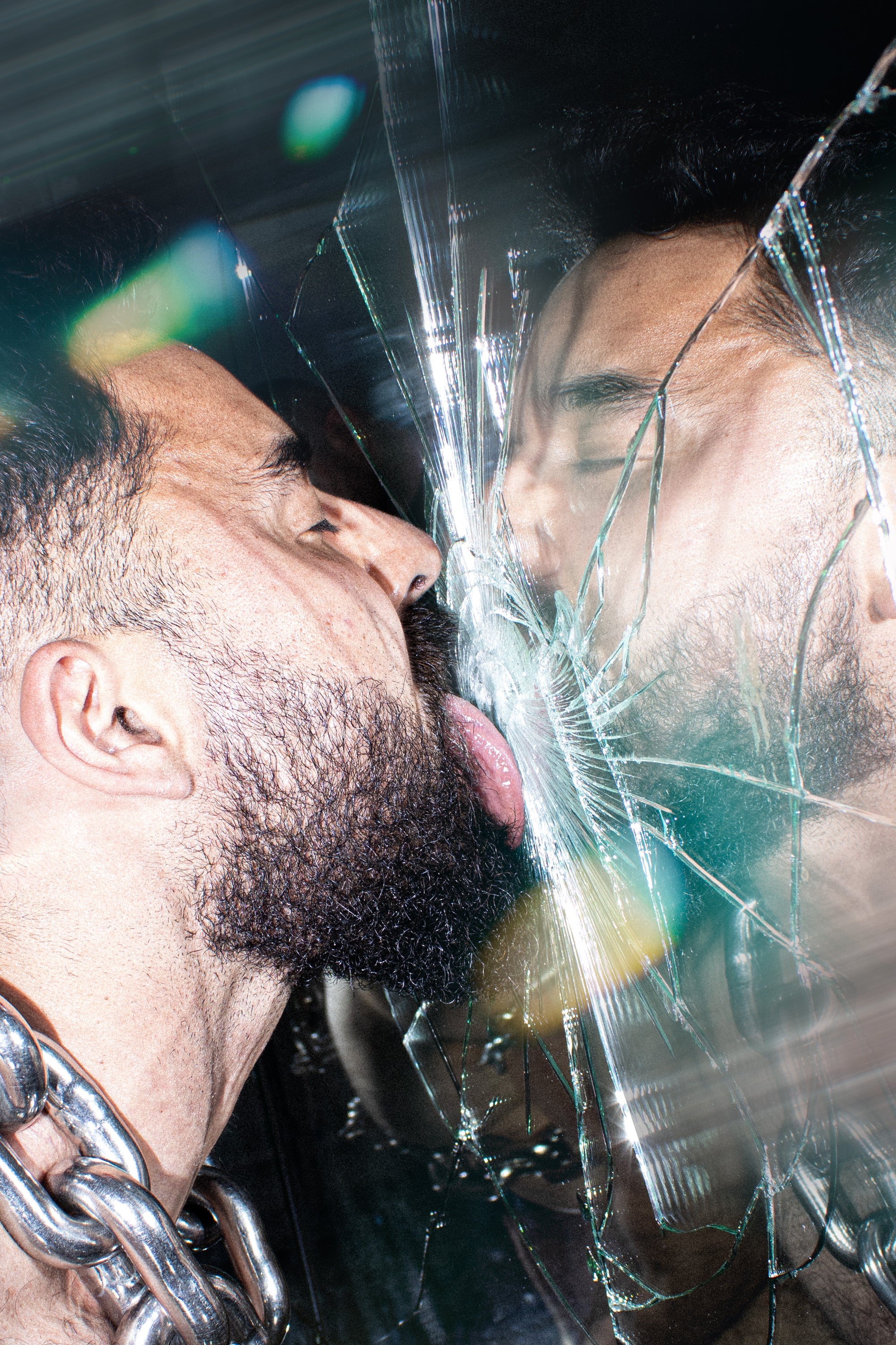 A close-up artistic photograph of a bearded man with a chunky metal chain collar, touching his reflection behind a heavily cracked glass surface.